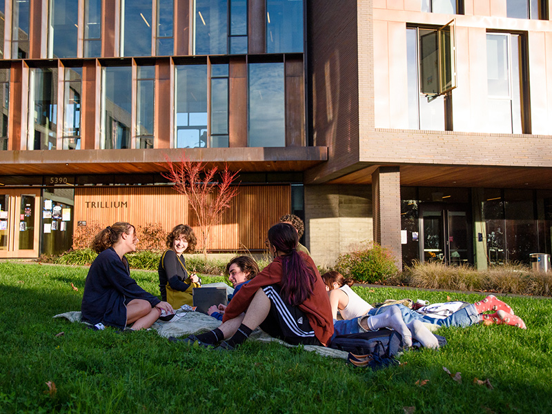 Six smiling and relaxed Reed students sit on a blanket outside Trillium, a residence hall