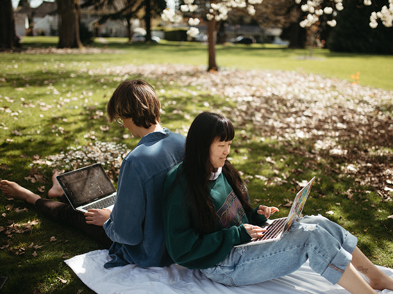 Two Reed students are typing on their laptops while sitting back-to-back in a shady spot under a tree