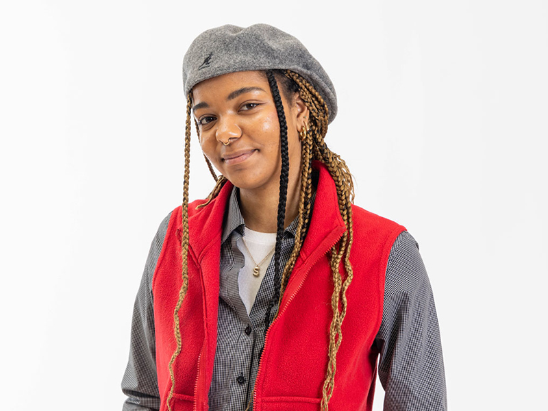 A portrait of Safi Zenger, who is wearing a red vest and has long braids under a gray beret.