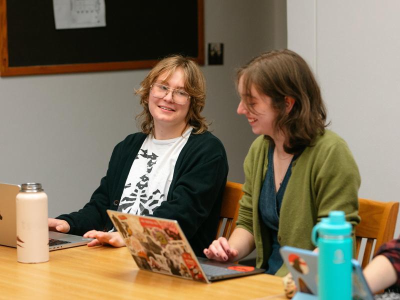 Two students sit side-by-side in a classroom at a conference table. They are talking and smiling.