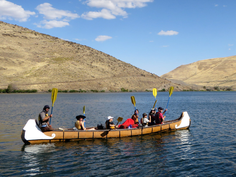 Six students and a guide float in a canoe on a stretch of water with two hills in the distance.