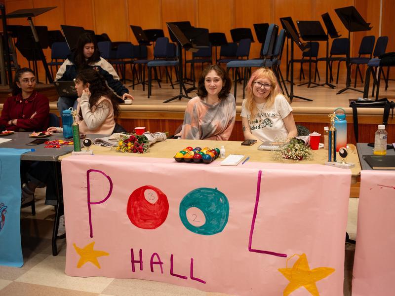 Two students sit at a table during an activities fair with a sign that says "pool hall." The two "o's" are represented by pool balls.