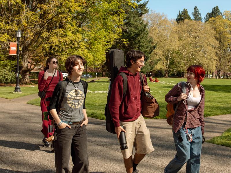 Four students walk in a group on a spring day with the great lawn stretching out behind them toward the edge of campus.