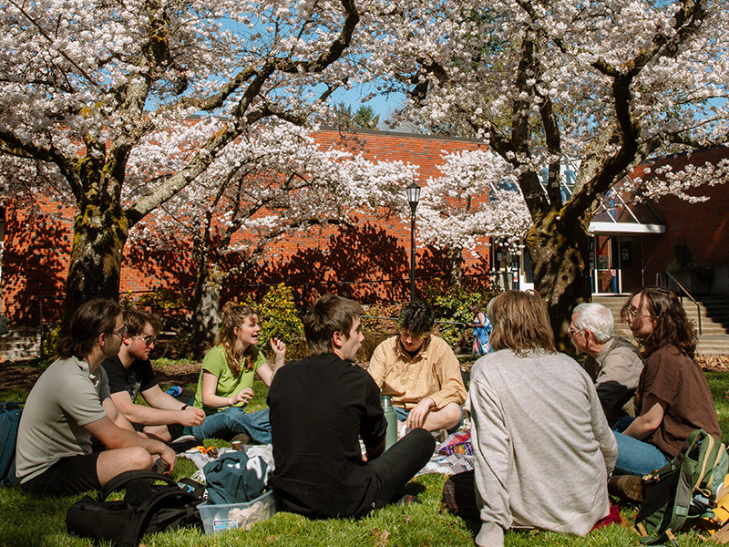 A group of students enjoying an outdoor classroom setting, sitting in a circle on the grass within Eliot Circle among cherry blossom trees in full bloom on a sunny afternoon.