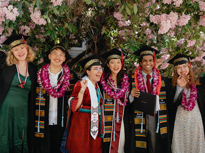 Six Reed College graduates smiling for the camera while wearing graduation caps, gowns, stoles, and floral leis.